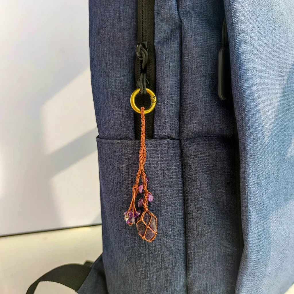 Violet Thread raw amethyst keychain used as a bag charm clipped to a dark backpack zipper. Demonstrates use as a backpack accessory.