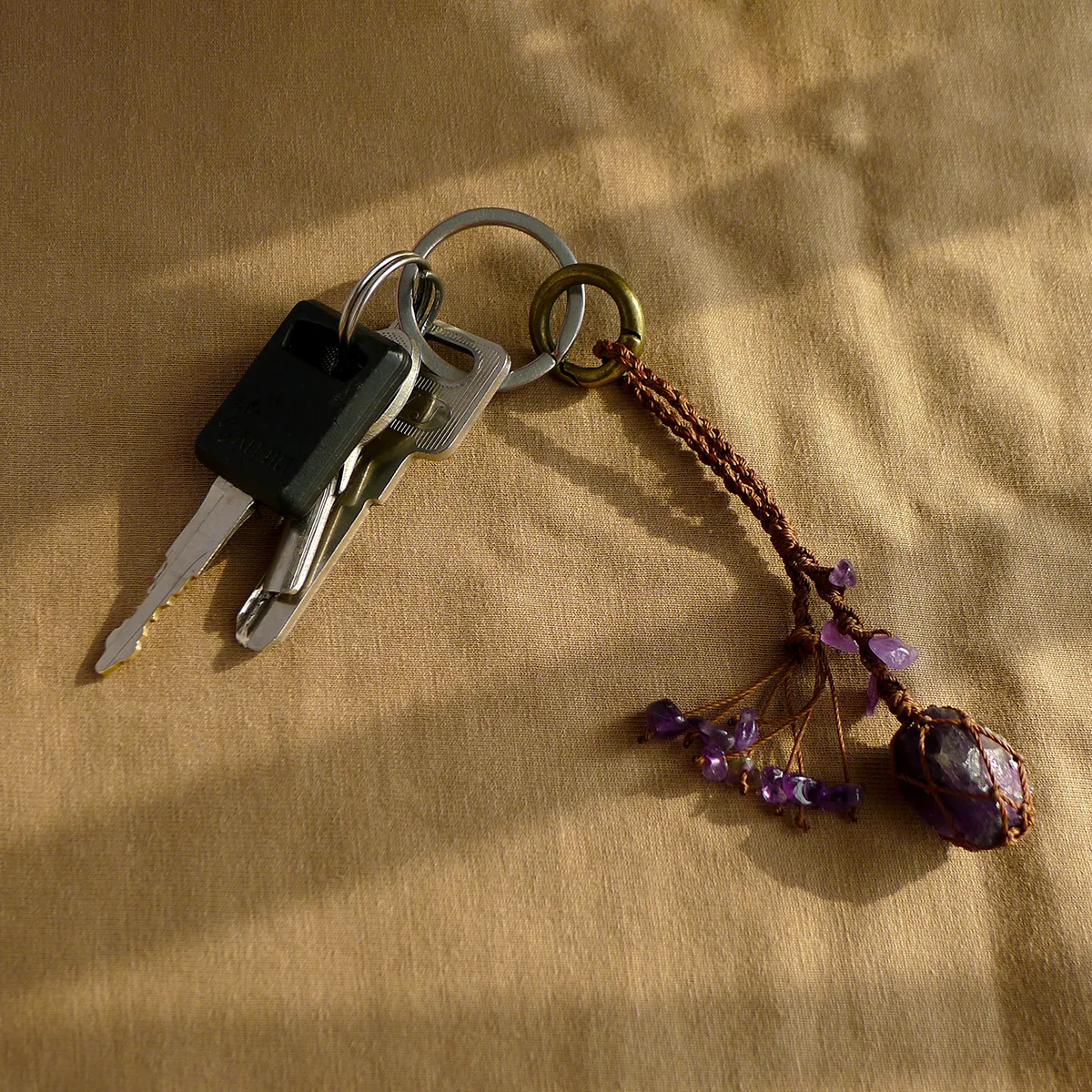 Violet Thread raw amethyst keychain clipped onto a keyring next to other everyday keys on tan fabric. Shows practical use as a keychain accessory.